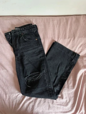Svarta raka jeans med slitning - Säljer ett par svarta jeans med rak passform och en snygg sliten detalj vid knät. Jeansen har klassisk femficksdesign och är tillverkade i denim. Perfekta för dig som gillar en avslappnad och trendig stil.