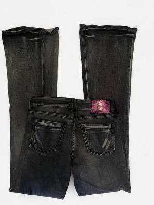 Svarta bootcut jeans med tvättad look - Säljer ett par svarta bootcut jeans med tvättad finish och klassiska fem fickor. Jeansen har en cool lapp bak i lila och svart och är tillverkade i ett stretchigt denimtyg. Perfekta för dig som gillar en avslappnad men trendig stil.