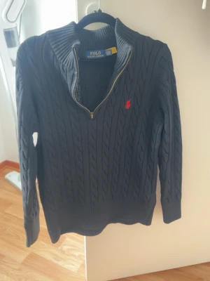 Svart kabelstickad tröja Polo Ralph Lauren - Kabelstickad svart tröja från Polo Ralph Lauren med halv dragkedja och röd broderad logga på bröstet. Tröjan har ribbade muddar och hög krage. Perfekt för dig som gillar klassisk och stilren design.