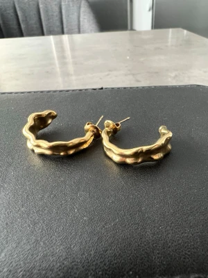 Guldpläterade vågiga creoler - Säljer ett par unika örhängen i guldpläterad metall med vågig, organisk form. Creolmodellen ger en cool och trendig look som sticker ut. Perfekta för dig som gillar statement-smycken och vill addera något extra till din stil. Köpta på Raglady. Aldrig använda 