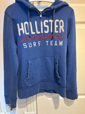 Blå Hollister hoodie med tryck - Blå hoodie från Hollister med vit broderad text 'HOLLISTER SURF TEAM' och röda vågor på bröstet. Hoodien har huva med dragsko, halv dragkedja och en stor magficka. Mjuk bomullsblandning och ribbade muddar vid ärmar och nederkant.