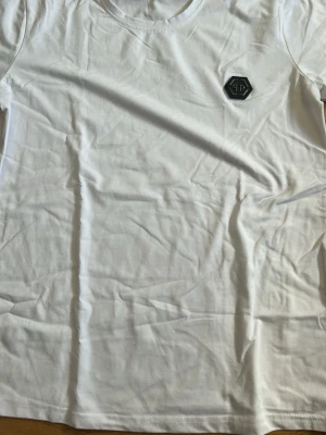 Vit t-shirt med PP-logga - Snygg vit t-shirt med rund hals och en svart PP-logga på bröstet. Tillverkad i mjuk bomull som känns skön mot huden. Perfekt basplagg för alla tillfällen och enkel att matcha med andra kläder.