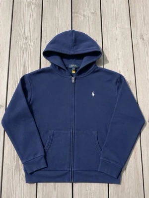 Marinblå hoodie från Polo Ralph Lauren - Klassisk marinblå hoodie från Polo Ralph Lauren med dragkedja framtill och vit broderad logga på bröstet. Hoodien har kängurufickor och ribbade muddar vid ärmar och nederkant. Tillverkad i mjuk bomullsblandning för skön komfort.