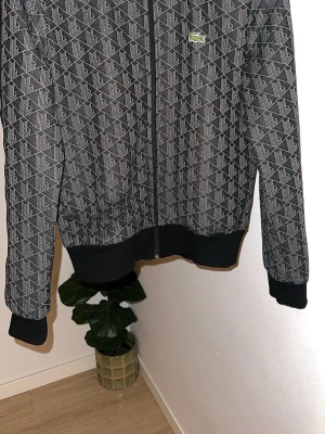 Svart och grå hoodie med mönster - Snygg svart och grå hoodie med hel dragkedja och geometriskt mönster. Tröjan har ribbade muddar och nederkant samt en liten broderad logga på bröstet. Perfekt för dig som gillar streetwear och vill sticka ut lite extra.