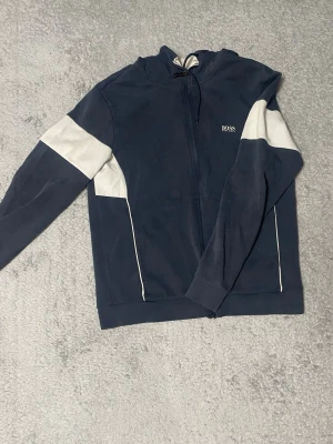 Mörkblå hoodie med vita detaljer - Snygg mörkblå hoodie från BOSS med vita paneler på ärmarna och vita detaljer längs sidorna. Dragkedja framtill, huva med dragsko och diskret BOSS-logga på bröstet. Perfekt för en avslappnad och sportig stil.