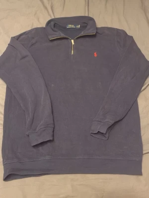 Marinblå halvzip från Polo Ralph Lauren - Snygg marinblå tröja med halv dragkedja från Polo Ralph Lauren. Klassisk röd logga broderad på bröstet och ribbade muddar vid ärmslut och nederkant. Perfekt till jeans eller chinos för en clean look.
