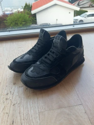 Svarta sneakers från Valentino - Snygga svarta sneakers från Valentino med coola camo-detaljer och klassisk låg siluett. Skorna har en mix av mocka och läder, samt svarta skosnören och gummisula. Perfekta för dig som vill ha en stilren men ändå unik look.