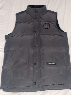 Grå dunväst från Canada Goose - Snygg grå dunväst från Canada Goose med hög krage och tryckknappar framtill. Västen har ett broderat svartvitt märke på bröstet och en diskret logga på ena fickan. Perfekt för lager-på-lager och håller dig varm under kyliga dagar.