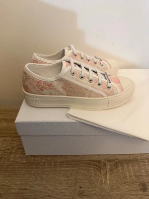 Dior rosa låga sneakers - Snygga låga sneakers från Dior i ljusrosa och vitt med broderat mönster. Skorna har vita snören, gummisula och detaljer med Dior-logga på sidan och tungan. Perfekta för dig som vill ha en exklusiv och trendig look.