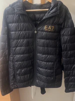 Svart pufferjacka från EA7 Emporio Armani - Snygg svart pufferjacka från EA7 Emporio Armani med guldfärgad logga på bröstet. Jackan har dragkedja framtill, två sidofickor med dragkedja och en hög krage. Tillverkad i lätt, quiltat material som håller värmen under kalla dagar.