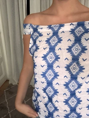Blåvit offshoulder topp med mönster - Säljer en blåvit offshoulder topp med grafiskt mönster. Toppen kan även bäras som ärmlös och har en lös passform. Perfekt till sommaren och tillverkad i ett lätt bomullstyg.