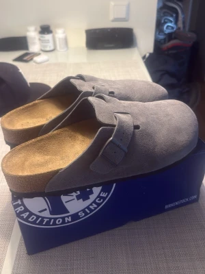Birkenstock Boston - Säljer ett par gråa tofflor från Birkenstock i mjuk mocka med klassisk design och justerbart spänne. Innersulan är i kork för extra komfort och fotriktighet. Perfekta för dig som vill ha både stil och bekvämlighet hemma.