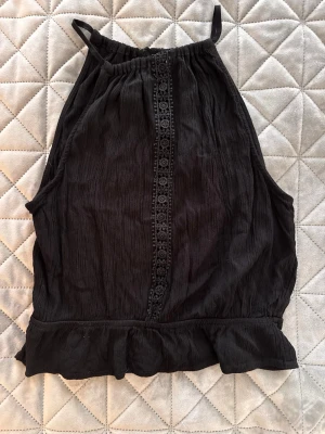 Svart linnetopp med spetsdetalj - Säljer en svart linnetopp med tunna axelband och dekorativ spets längs mitten framtill. Toppen har volangkant nedtill och är tillverkad i ett lätt, skrynkligt tyg som ger en avslappnad känsla. Perfekt för varma dagar eller att styla lager på lager.