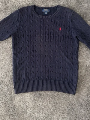 Mörkblå kabelstickad tröja Polo Ralph Lauren - Kabelstickad mörkblå tröja från Polo Ralph Lauren med rund halsringning och röd broderad logga på bröstet. Tröjan har ribbade muddar vid ärmslut och nederkant. Klassisk och stilren design som passar till mycket. Storlek 14-16 men ganska liten i storlek. Passar dig runt 164cm.