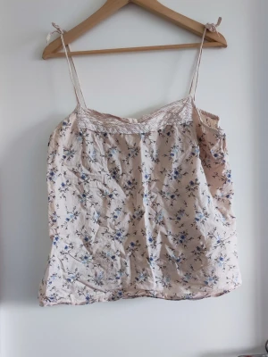 Blommig beige linnetopp med spets - Söt beige linnetopp med tunna axelband och blommigt mönster i blått och lila. Toppen har spetsdetaljer upptill och är perfekt för varma dagar. Lätt och luftig känsla med en feminin touch.