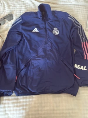 Blå Real Madrid träningströja Adidas - Snygg blå träningströja från Adidas med Real Madrid-logga på bröstet. Tröjan har halv dragkedja, vita Adidas-ränder på ärmarna och rosa detaljer. Tillverkad i ett funktionsmaterial som passar perfekt till träning eller uppvärmning.