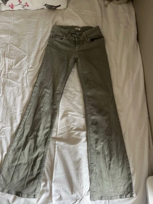 gröna bootcut jeans med låg midja - Säljer ett par gröna bootcut jeans med låg midja och dubbla knappar i linningen. Jeansen har klassiska fem fickor och bootcut ben som ger en snygg retrovibe. Perfekta för dig som gillar en avslappnad men trendig stil.