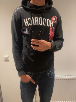 Svart hoodie från Hoodrich - Svart hoodie från Hoodrich med vita och röda detaljer, broderade patches och stor ficka framtill. Klassisk huva med dragsko och ribbade muddar. Perfekt för en streetwear-look.