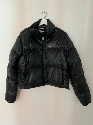 Svart pufferjacka från Columbia - Snygg svart pufferjacka från Columbia med hög krage och dragkedja framtill. Jackan har en boxig passform och är tillverkad i glansigt material som ger en modern look. Perfekt för kalla vinterdagar och riktigt varm.