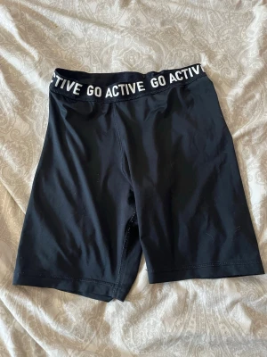Svarta cykelshorts från Kappahl - Svarta cykelshorts från Kappahl med bred resår i midjan där det står 'GO ACTIVE' i vitt. Shortsen är tillverkade i stretchigt syntetmaterial och har en tight passform som passar perfekt till träning eller chill.🌸