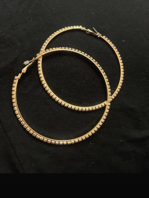 Stora guldfärgade hoops örhängen - Säljer ett par stora, runda hoops i guldfärg med fyrkantiga detaljer längs hela ringen. Örhängena har en klassisk klickstängning och är perfekta för dig som vill ha ett statement-smycke. Materialet ser ut att vara metall med guldfinish.