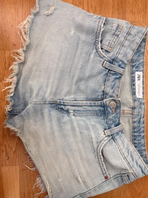 Ljusblå jeansshorts från Zara - Säljer ett par ljusblå low waist jeansshorts från Zara med fransiga kanter och slitna detaljer. Klassisk femficksmodell med knappgylf och bälteshällor. Perfekta för varma dagar och ger en avslappnad vibe.