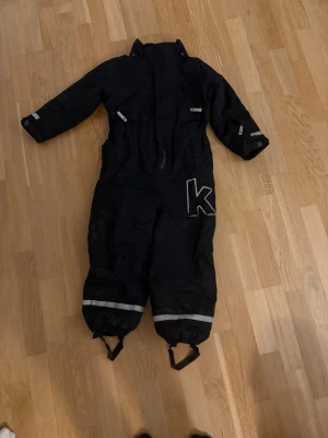 Svart overall med reflexdetaljer - Mörkblå täckbyxa i overallmodell med långa ärmar och ben, reflexdetaljer på armar och ben samt elastiska fotband. Stor K-logga på ena benet. Perfekt för kalla dagar och utomhusaktiviteter.