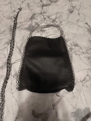Svart handväska med kedjedetaljer - Snygg svart handväska i konstläder med silverfärgade kedjor längs kanterna och som axelrem. Väskan har en mjuk, något slouchy form och en cool, modern look. Perfekt för dig som gillar edgy accessoarer och vill sticka ut.
