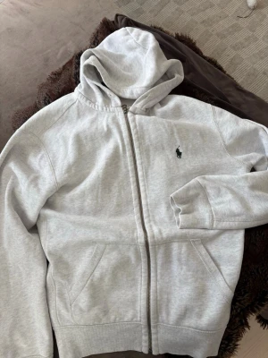  Polo Ralph Lauren Hoodie  - Säljer en ljusgrå hoodie från Polo Ralph Lauren med dragkedja och huva. Klassisk logga broderad på bröstet och två fickor framtill. Tillverkad i mjukt bomullsmaterial med ribbade muddar vid ärmslut och nederkant. Perfekt för en chill och stilren look.