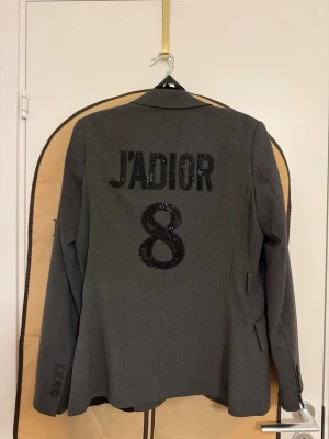 Grå kavaj  - Snygg grå kavaj med svart broderad siffran 8 på ryggen. På sidan finns en liten broderad bi och initialerna CD. Kavajen har klassisk passform, knappar vid ärmslut och fickor fram. Perfekt för dig som vill sticka ut med coola detaljer.