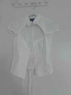Vit kortärmad blus med knytband - Stilren vit blus med korta puffärmar och knytband framtill. Klassisk krage och lätt figursydd passform. Perfekt att matcha med jeans eller kjol för en trendig look. Tillverkad i ett tunt, vävt material.