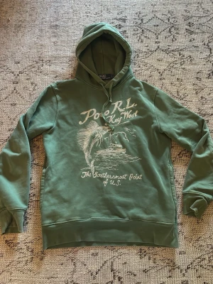 Grön hoodie från Polo Ralph Lauren - Snygg grön hoodie från Polo Ralph Lauren med tryck på bröstet som visar en fisk och texten 'Key West'. Hoodien har dragsko i huvan och ribbade muddar vid ärmar och nederkant. Perfekt för en avslappnad stil och riktigt skön att bära. Archive
