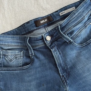 Blå Replay Anbass jeans - Snygga blå Replay Anbass jeans med klassisk femficksdesign och diskreta slitningar. Jeansen har en rak passform och är tillverkade i mjukt denimtyg. Märkesdetaljer syns på bakfickan och patchen i midjan. Perfekta för en avslappnad och trendig look.