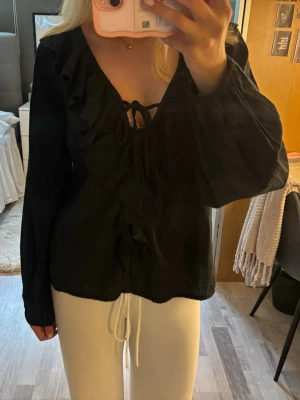 Svart blus  - Säljer en svart blus med djup v-ringning, volangdetaljer längs urringningen och snygg knytning framtill. Blusen är från Lager 157 i storlek S❣️knappt använd!