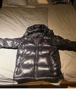 Ralph lauren puff jacka svart - Snygg svart pufferjacka från Polo Ralph Lauren med blank finish och huva. Jackan har dragkedja framtill, två sidofickor med dragkedja och en röd logga på bröstet. Perfekt för kalla vinterdagar och riktigt trendig look.