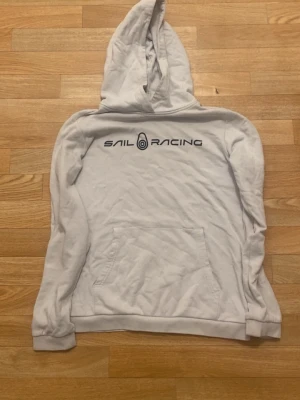 Vit hoodie från Sail Racing - Säljer en vit hoodie från Sail Racing med svart logga och text på bröstet samt en mindre logga på huvan. Hoodien har en klassisk känguruficka framtill och ribbade muddar vid ärmslut och nederkant. Perfekt för en avslappnad och sportig stil.