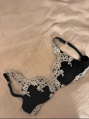 Svart push-up bh med spetsdetaljer - Snygg svart push-up bh med beige spetsdetaljer och broderier framtill. Bh:n har vadderade kupor och justerbara axelband. Elegant design med fina spetskanter som ger en lyxig känsla.