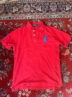 Röd pikétröja från Polo Ralph Lauren - Klassisk röd pikétröja från Polo Ralph Lauren med blå broderad logga på bröstet och två knappar vid kragen. Tröjan har korta ärmar och en bekväm passform, perfekt till jeans eller shorts. Ikonisk stil som aldrig går ur tiden.