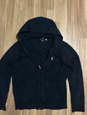 Svart hoodie från Polo Ralph Lauren - Snygg svart hoodie med dragkedja från Polo Ralph Lauren. Klassisk design med vit broderad logga på bröstet, två fickor framtill och justerbar huva med snören. Tillverkad i mjukt bomullsmaterial som är skönt att bära.