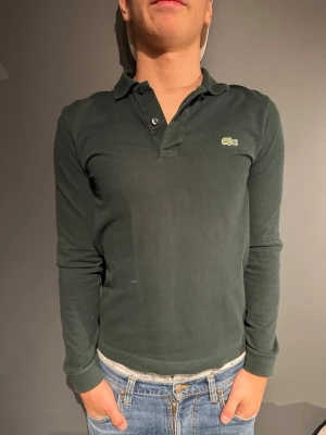 Mörkgrön långärmad piké Lacoste - Mörkgrön långärmad pikétröja från Lacoste med klassisk krage och knappslå framtill. Tröjan har den ikoniska krokodilloggan broderad på bröstet och är tillverkad i ett mjukt bomullsmaterial. Perfekt till jeans eller chinos. Modellen är 175cm och väger 70kg