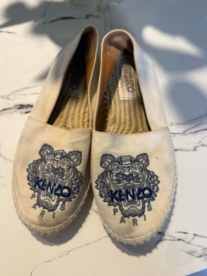 Beige espadriller med Kenzo-tryck - Säljer ett par beige espadriller från Kenzo med ikoniskt blått tigermotiv och texten 'KENZO PARIS' broderat på ovandelen. Skorna har klassisk flätad sula i jute och ovandel i canvas. Perfekta för dig som vill ha en cool och trendig look.