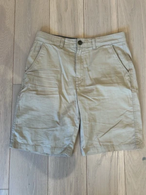 Beige bomullsshorts med fickor - Snygga beige shorts i bomull med raka ben och två sidofickor. Klassisk design med knapp och dragkedja framtill samt hällor för bälte. Perfekta för sommardagar och enkel att matcha med olika stilar.