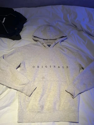 Jack & Jones hoodie - Snygg ljusgrå hoodie med texten 'ORIGINALS' tryckt över bröstet. Klassisk modell med huva och ribbade muddar vid ärmar och nederkant. Perfekt för en chill och avslappnad stil. Mjuk och skön bomullsblandning.