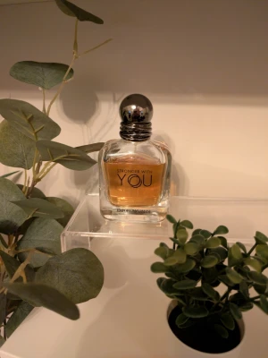 Stronger With You parfymflaska - Stronger With You från Emporio Armani är en elegant parfym i en genomskinlig glasflaska med rundade kanter och en silverfärgad, klotformad kork. Doften har en varm bärnstensfärgad vätska som syns tydligt genom flaskan.