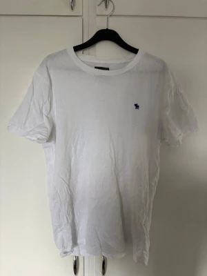 Vit t-shirt med blå logga - Klassisk vit t-shirt i bomull med rund hals och korta ärmar. På bröstet finns en liten blå broderad logga som ger en stilren touch. Perfekt basplagg till garderoben och enkel att matcha med allt. Bra skick fortfarande 