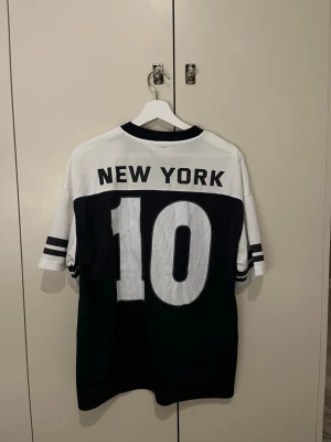 Svartvit jersey med New York-tryck - Snygg svartvit t-shirt med sportig look och stora siffran 10 på både fram- och baksidan samt texten NEW YORK på ryggen. T-shirten har korta ärmar med dubbla ränder och V-ringad hals. Perfekt för dig som gillar streetwear och sportig stil.