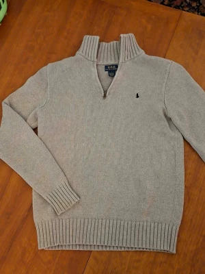 Ljusgrå stickad tröja Polo Ralph Lauren - Ljusgrå stickad tröja från Polo Ralph Lauren med halv dragkedja och hög krage. Tröjan har ribbade muddar vid ärmslut och nederkant samt en liten mörkblå logga broderad på bröstet. Perfekt för lager på lager och riktigt snygg till jeans. Storleken är Large i barn storlek, 14-16 år men använde när jag var 13-14 år.