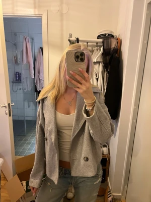 Ljusgrå kort kappa med bred krage - Säljer en ljusgrå kappa i kortare modell med bred krage och dubbelknäppning framtill. Aldrig använd ute.