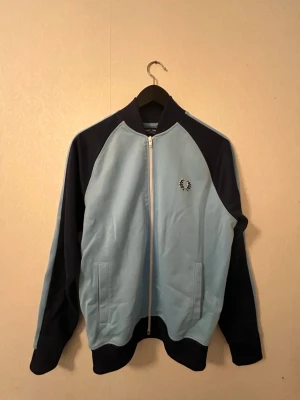 Fred Perry zip tröja - Snygg zip-tröja från Fred Perry i ljusblått och mörkblått med raglanärmar och broderad logga på bröstet. Tröjan har ribbade muddar och två fickor framtill. Perfekt för en sportig och avslappnad look.