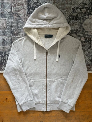 Grå hoodie från Polo Ralph Lauren - Snygg ljusgrå hoodie från Polo Ralph Lauren med dragkedja, huva med vita snören och två fickor framtill. Klassisk logga broderad på bröstet. Tillverkad i mjuk bomullsblandning och har ribbade muddar vid ärmar och nederkant. Perfekt för en chill och stilren look. Mycket liten slitning i huvan (se bild nr.7), men annars är tröjan i riktigt fint skick!